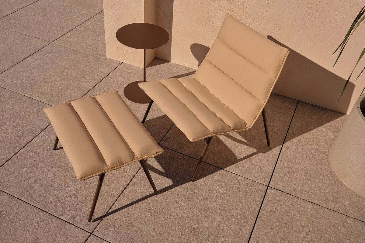Aria Outdoor Chair and Ottoman in a warm neutral finish, styled on a tiled outdoor terrace beside a small round side table.