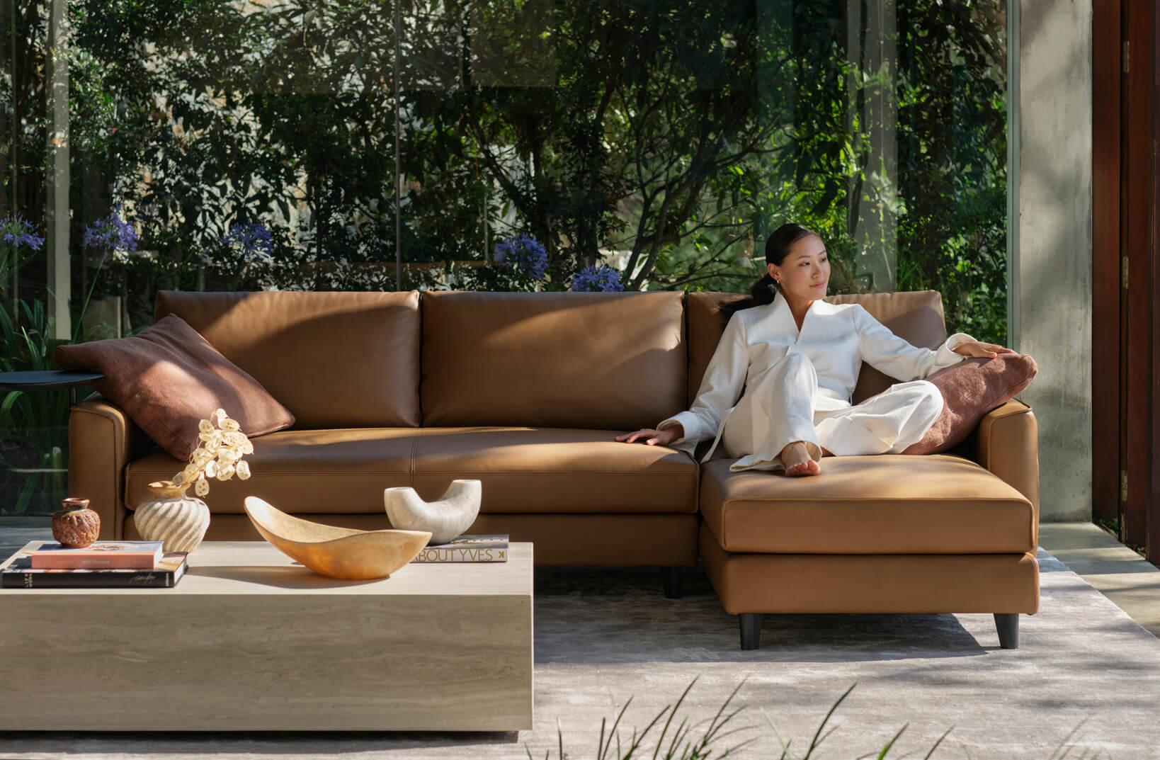 A woman lounging on a tan-leather sofa with chaise, in a biophilic-designed interior with greenery, natural textures and accessories.