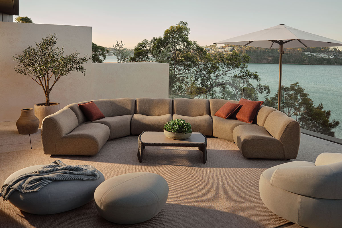 Light brown modular outdoor sofa with russet cushions, styled in an outdoor living space on a patio above a coastal bay.