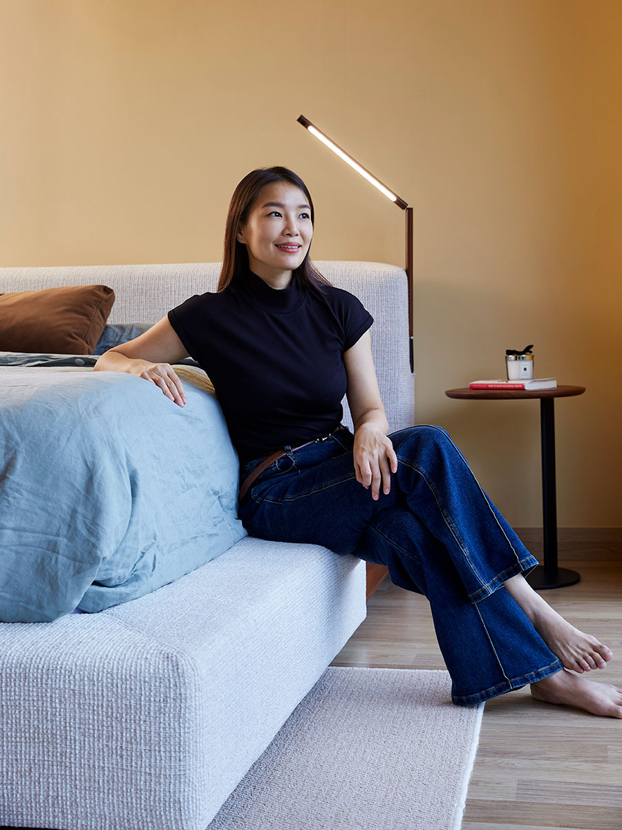 Person sitting on a modern upholstered bed with a minimalist lamp and side table in the background.